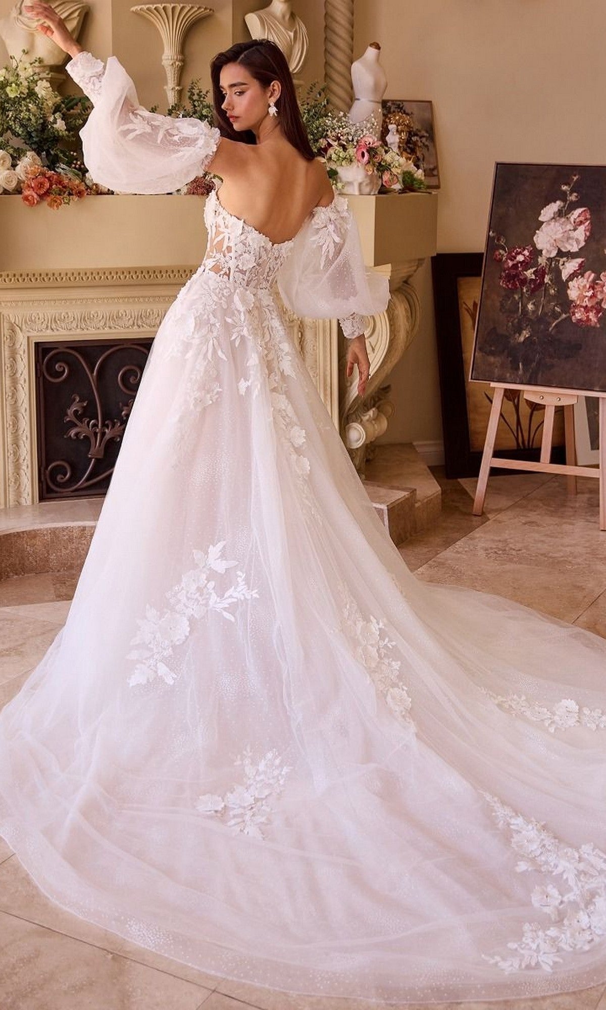 A woman wearing the Andrea and Leo WL018 Long Wedding Dress, featuring a strapless off-shoulder lace design with floral details and a long train, stands indoors near a fireplace and elegant floral decorations.